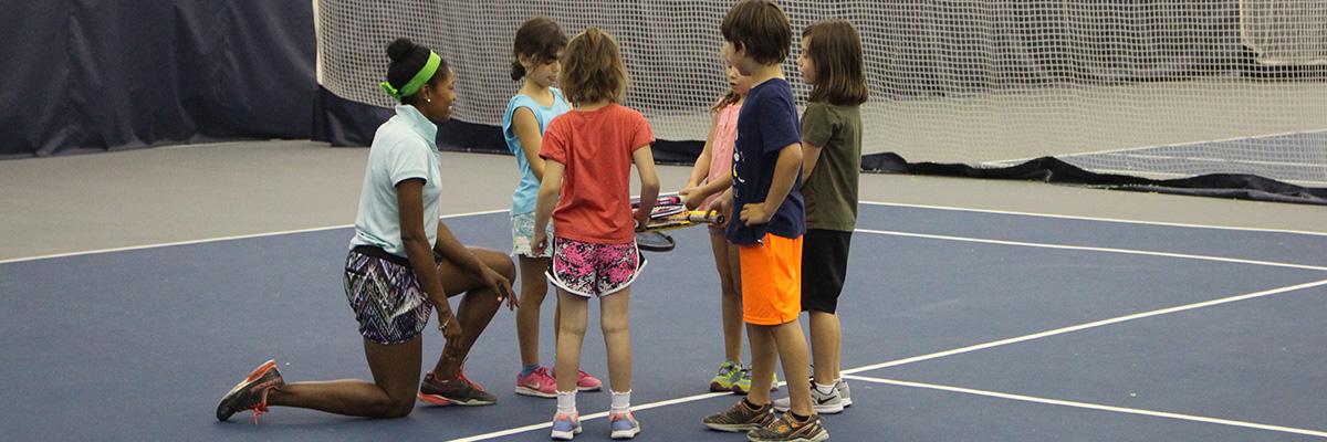 An instructor kneeling and talking to five children on a tennis court.